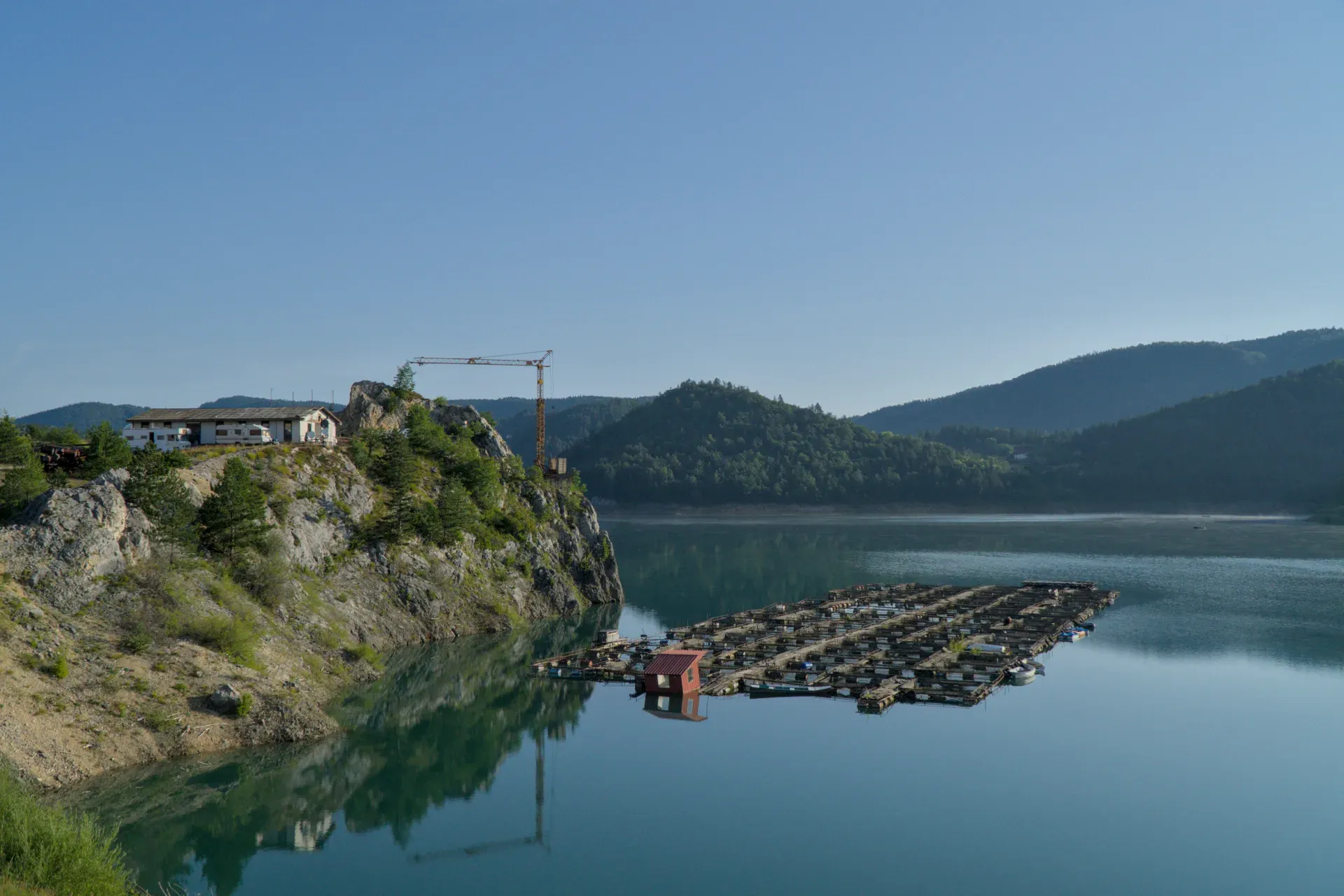 Izlazak sunca na jezeru Zaovine, Srbija. Mirna voda odražava planine i vedro nebo sa plutajućim strukturama.