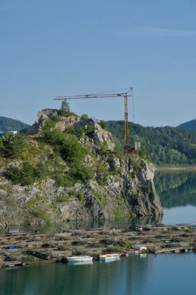 Vista matutina del Lago Zaovine con una grúa sobre una roca y piscifactorías flotantes en primer plano.