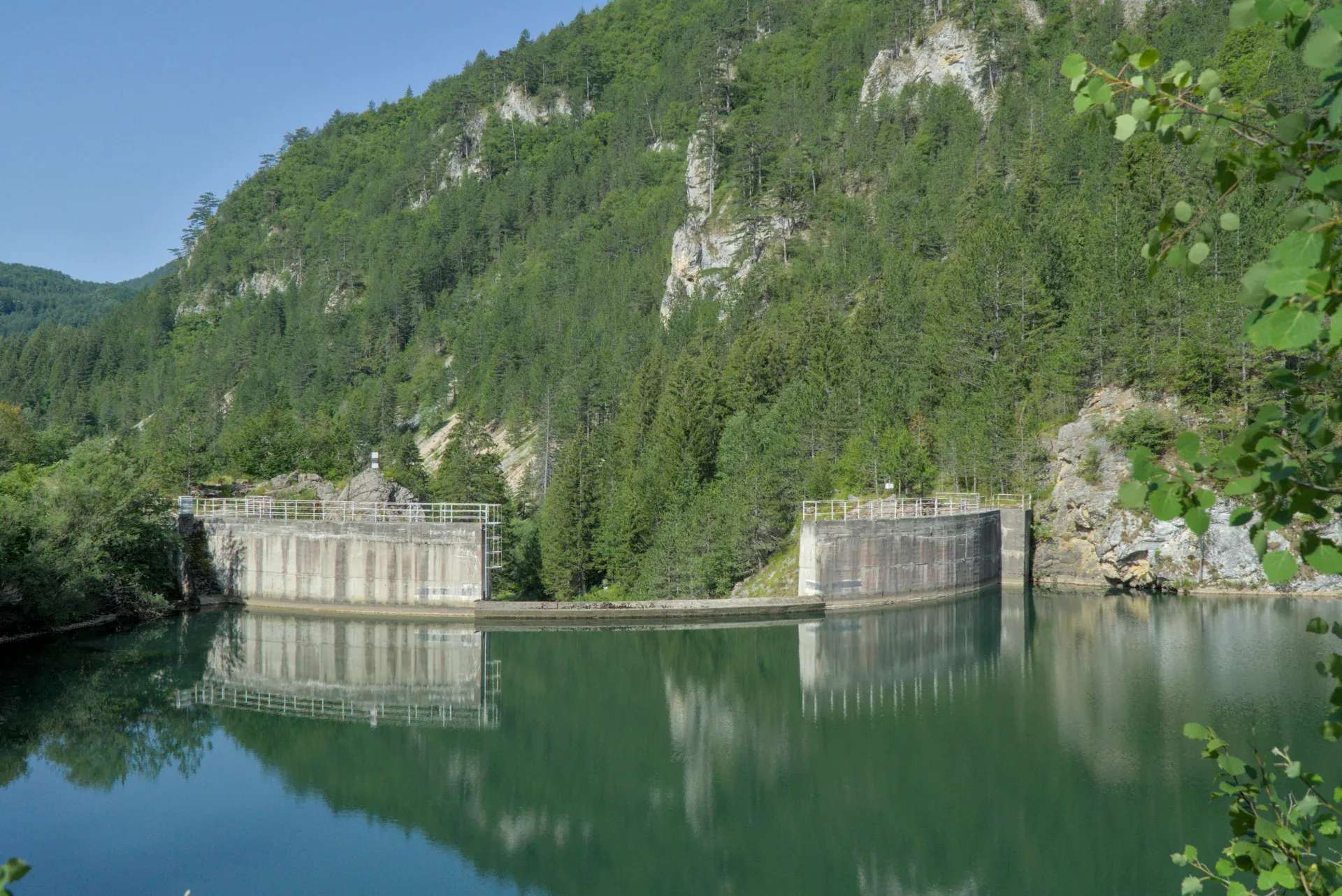 Il Lago Spajići con la sua diga in cemento, riflesso in acque verdi contro colline boscose.