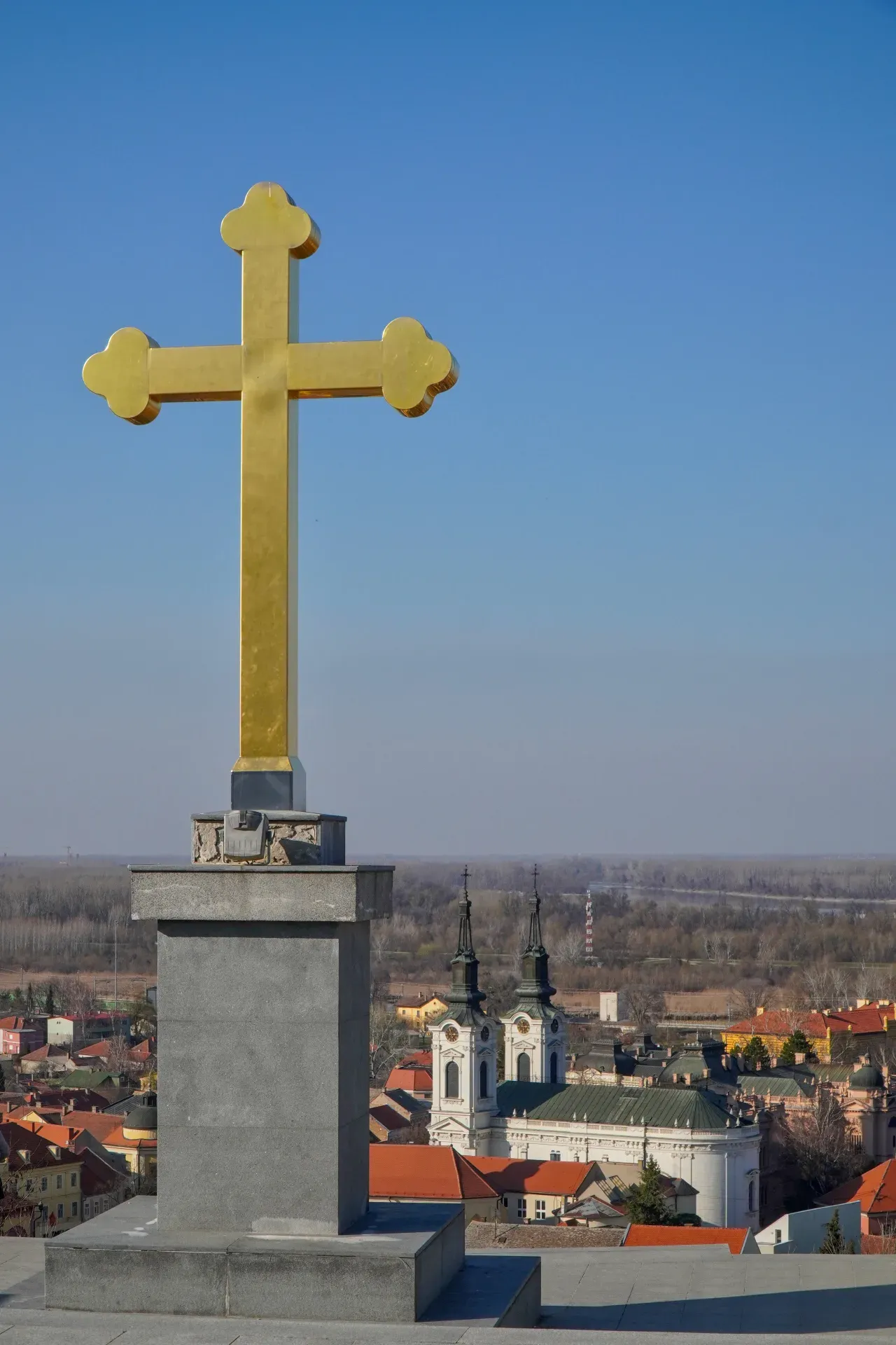 Une croix dorée proéminente se dresse sur un ciel bleu clair, surplombant une vue panoramique de Sremski Karlovci, Serbie.