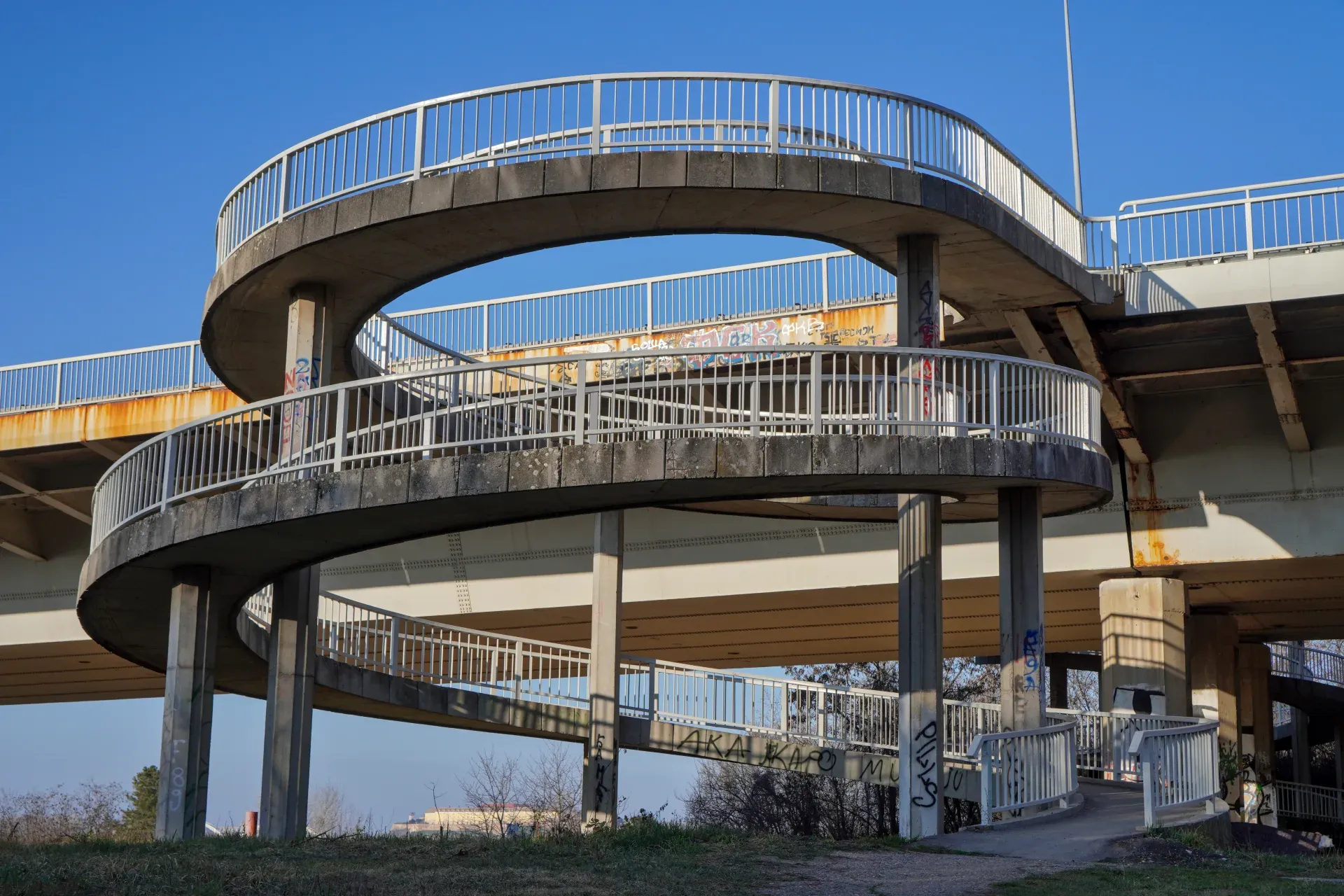 Une rampe en spirale de béton avec des garde-corps métalliques, descendant du Pont de la Liberté vers le Parc Kamenički.