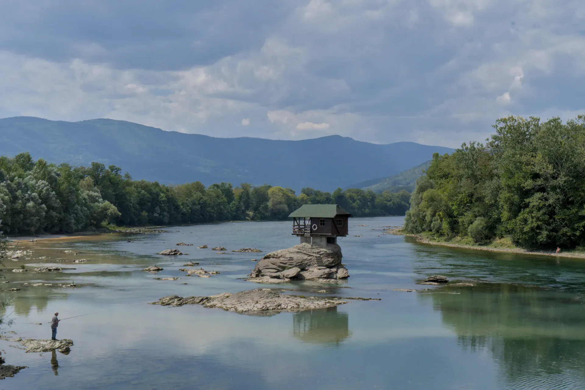 Una pequeña casa en una roca en el río Drina, con un pescador solitario lanzando una caña cerca.