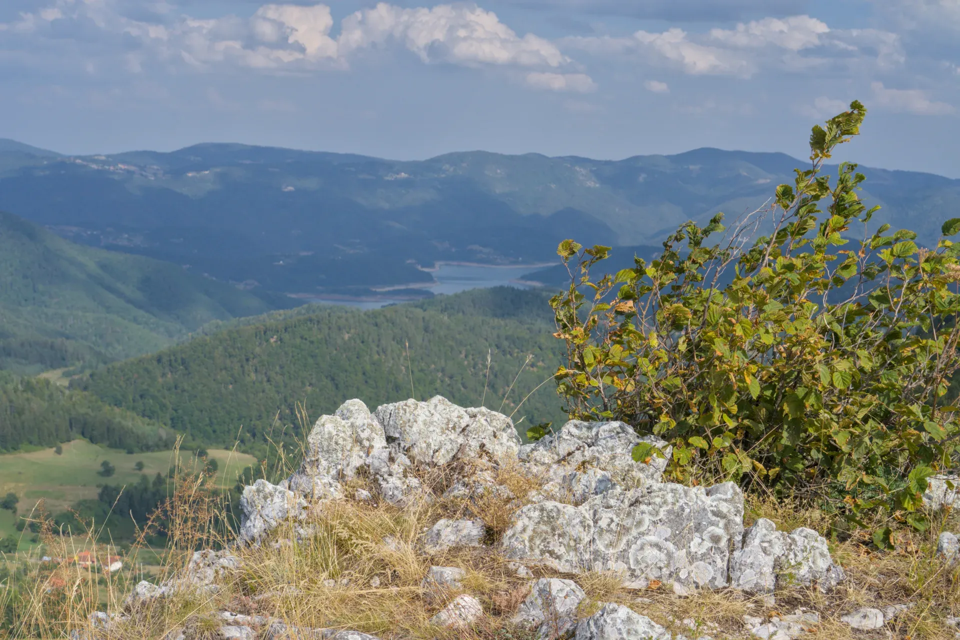 Lac Zaovine vu des montagnes Jankovica, Serbie, avec des rochers et de la végétation au premier plan et des collines ondulantes.