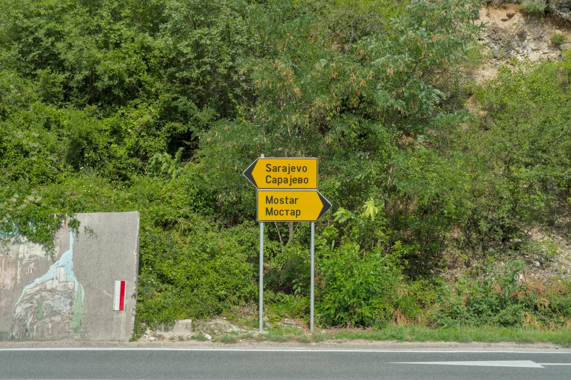 Wegweiser zeigt nach Mostar und Sarajevo, umgeben von grünem Gestrüpp, ein Zeichen für die Reise Route.