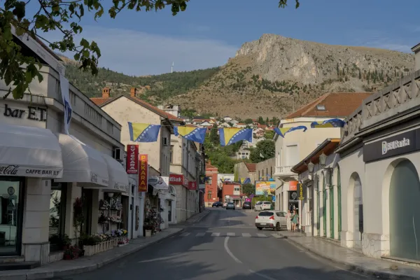 Straße in Mostar mit bosnischen Flaggen im Abendlicht, Berg im Hintergrund.