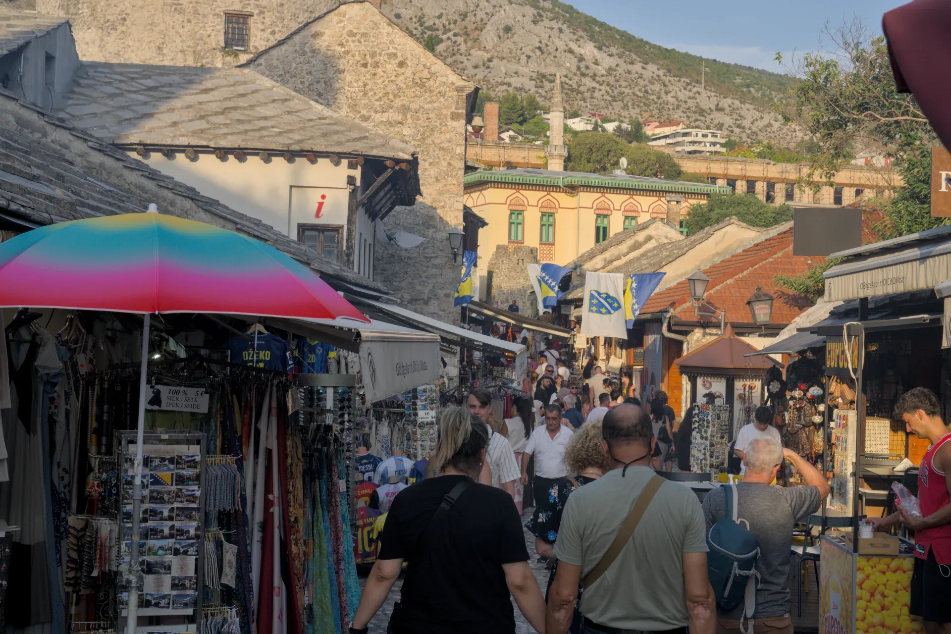 A bustling street in Mostar's Old Town at dusk, lined with shops and the backdrop of mountains.