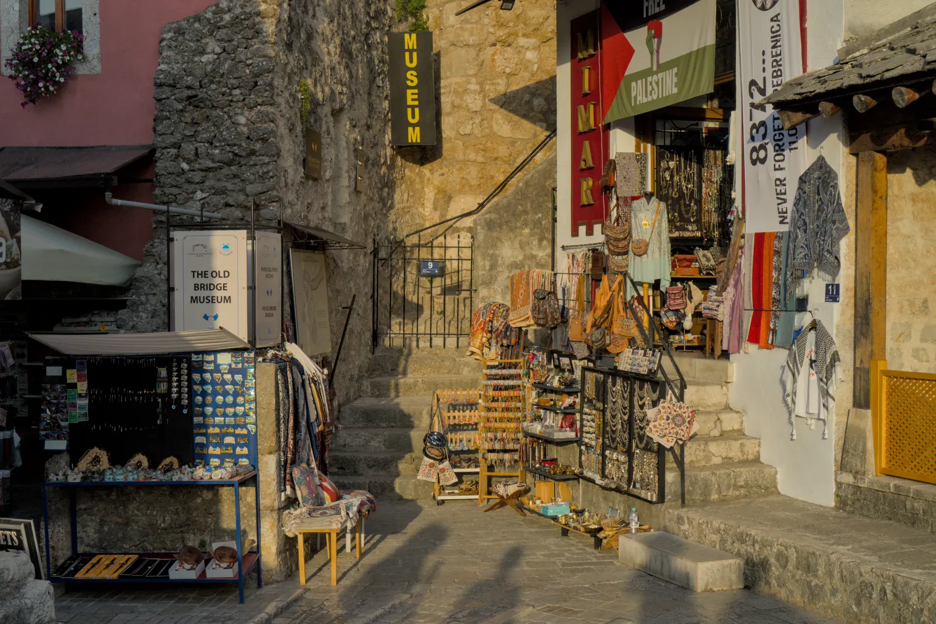 Rue atmosphérique de la vieille ville de Mostar durant l'heure dorée, avec des étals d'artisans et des bâtiments en pierre.
