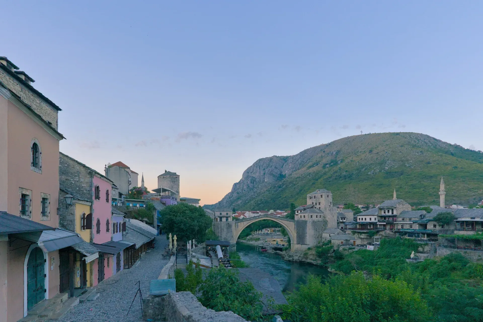 Panorama previo al amanecer del casco antiguo de Mostar con casas coloridas, el puente histórico y un río tranquilo.