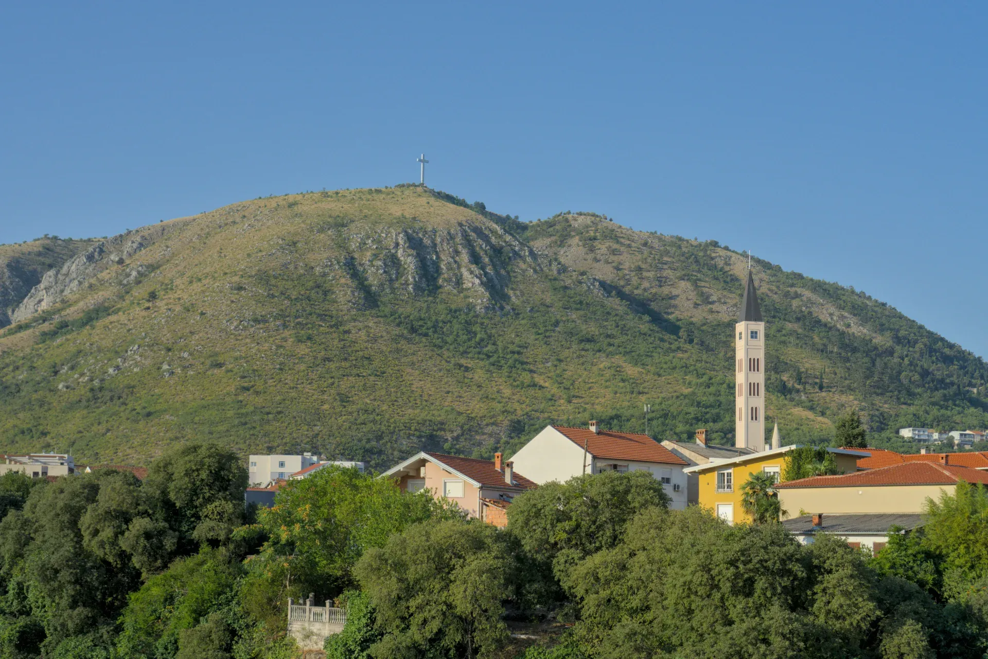 Eine weitläufige Landschaftsaufnahme von Mostar mit einem markanten Kreuz auf einem Berggipfel und einem schlanken Kirchturm im Vordergrund.