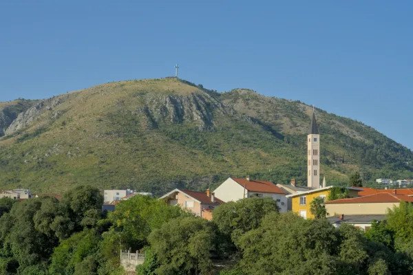Panoramski likovni pejzaž Mostara sa velikim krstom na brdu i vitkim crkvenim tornjem u prvom planu.