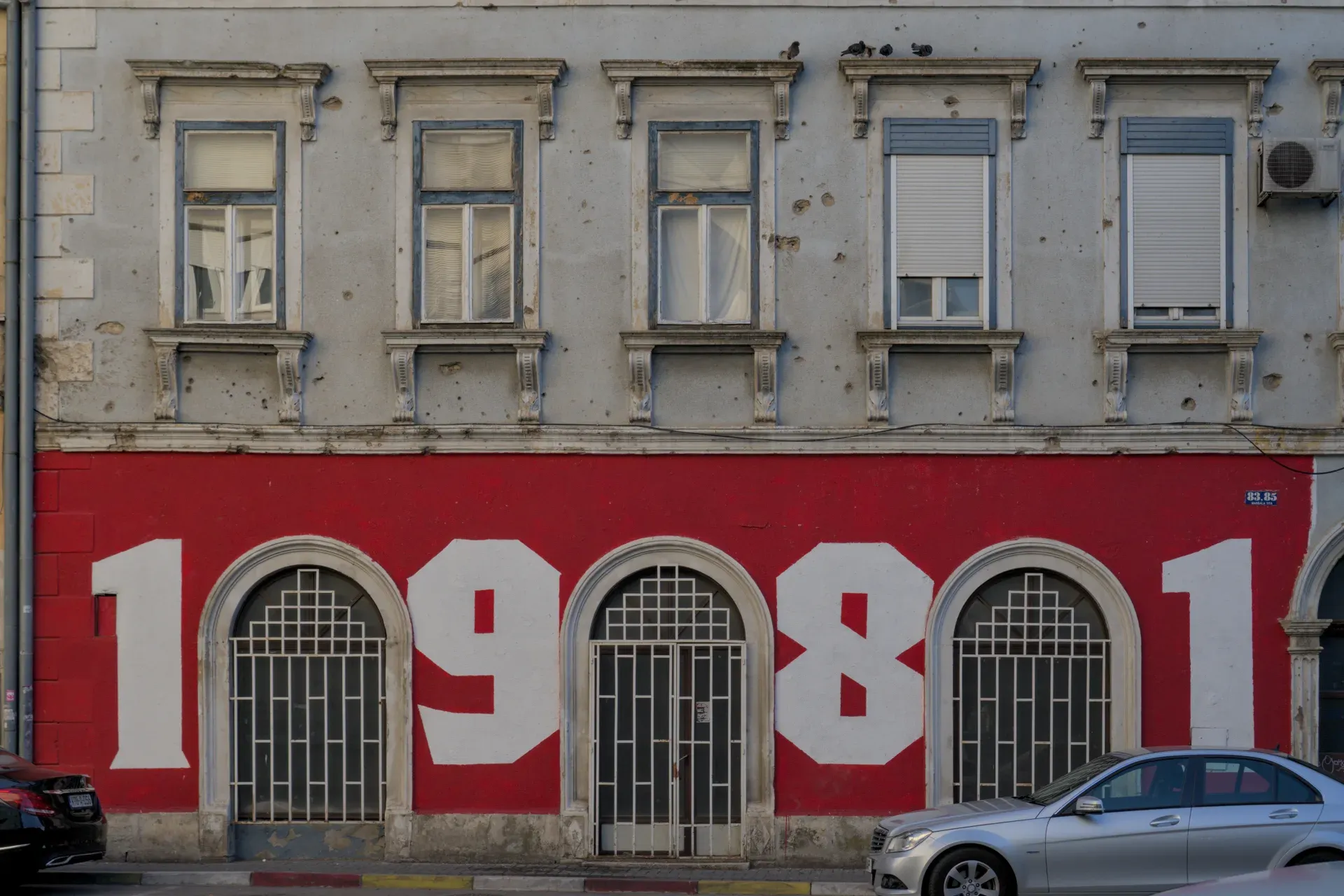 Graffiti "1981" on a red wall in Mostar, Bosnia and Herzegovina, with historical windows and facade details.