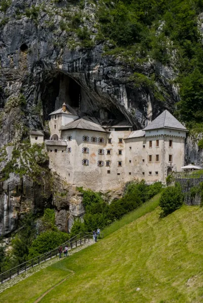 El Castillo de Predjama anidado en una pared de roca masiva, con follaje verde y visitantes en un camino debajo.