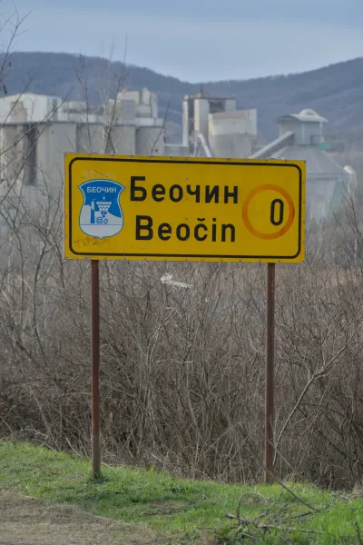 Señal de carretera amarilla para 'Beočin' en serbio y latín con una fábrica de cemento al fondo.