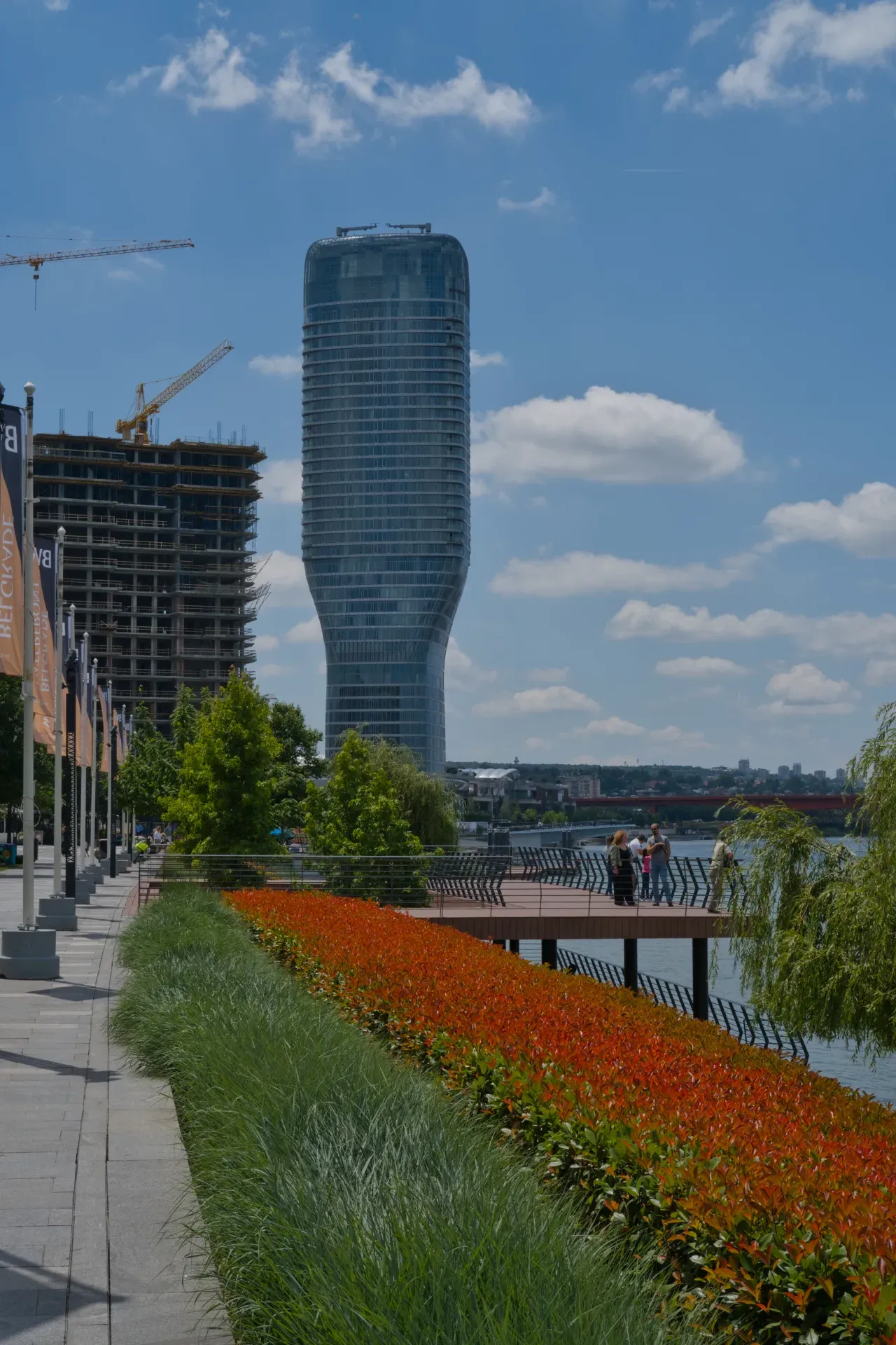 Modernes Gebäude am Fluss mit leuchtend roten Blumenbeeten und Menschen auf der Promenade in Belgrad.