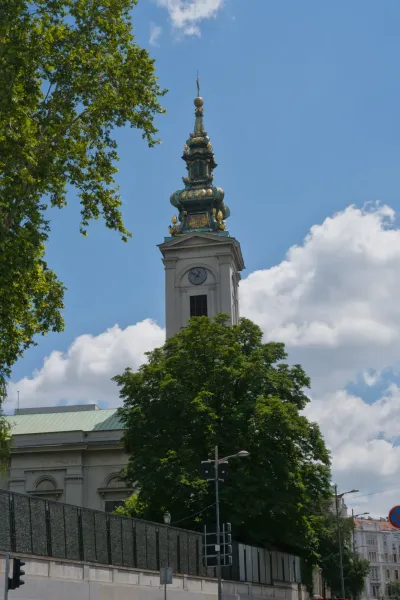 Elegante campanario de iglesia en Belgrado contra un cielo azul con nubes, enmarcado por árboles.