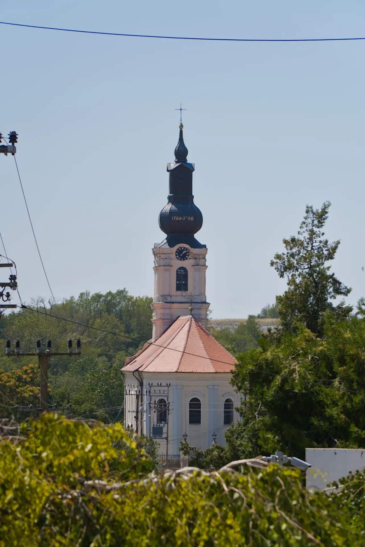 Fotografia ritratto di una chiesa a Irig, Serbia, con una torre e un tetto distintivi, su uno sfondo di cielo azzurro.
