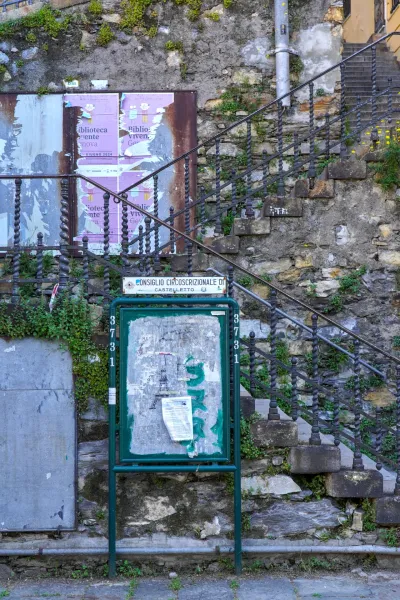 Una antigua escalera de piedra con una barandilla de metal ornamentada asciende junto a una pared de piedra. En primer plano, un tablón de anuncios verde muestra carteles desgastados y plantas exuberantes trepan por la pared y las escaleras.