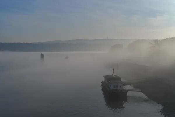 Pristanište na Dunavu u gustoj magli sa čamcem kod obale, stvarajući misterioznu atmosferu.