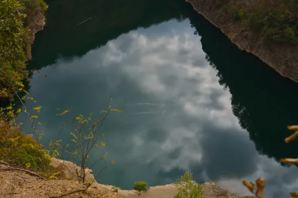Dve patke polako plivaju po zrcalnoj površini Ledinačkog jezera, odražavajući oblake. Pogled sa stene.