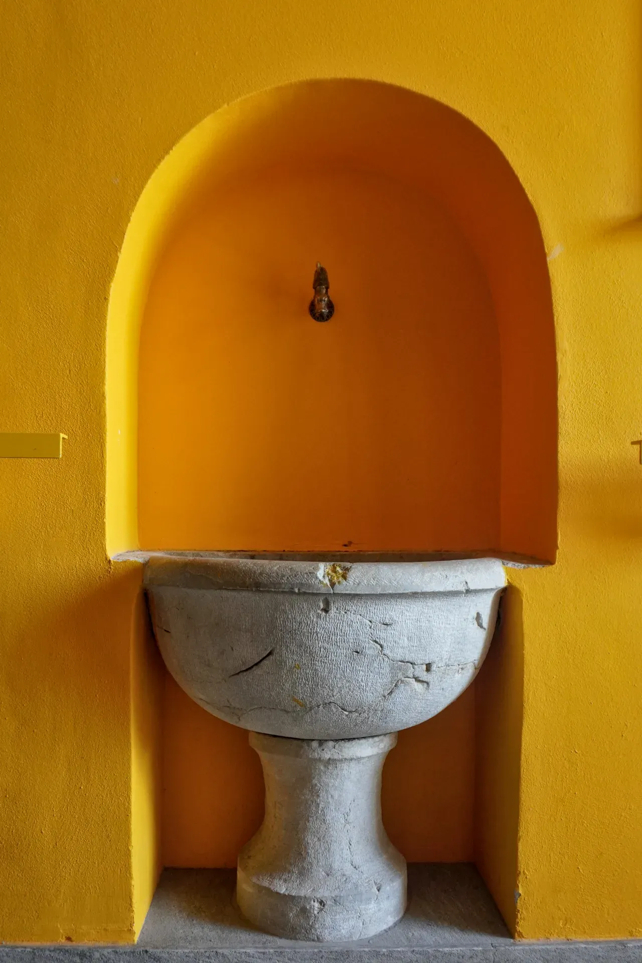 Eine Vintage-Steinspüle sitzt in einer leuchtend gelben bogenförmigen Nische mit einem kleinen Messingwasserhahn darüber, vor einer strukturierten gelben Wand. Der Boden ist mit gedämpften grauen Fliesen ausgelegt.