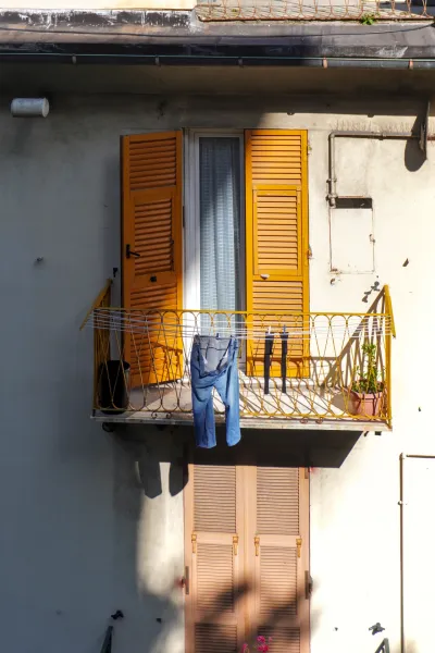 Un balcón en Génova, Italia, con contraventanas de color naranja brillante en la ventana, vaqueros azules y calcetines negros en un tendedero, y una pequeña planta verde en maceta.