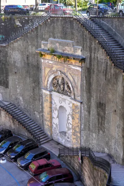 Un monumento histórico erosionado con un relieve escultural bajo un arco, sobre un muro de piedra rugosa, con numerosas escaleras exteriores y un aparcamiento con coches debajo en Génova, Italia.