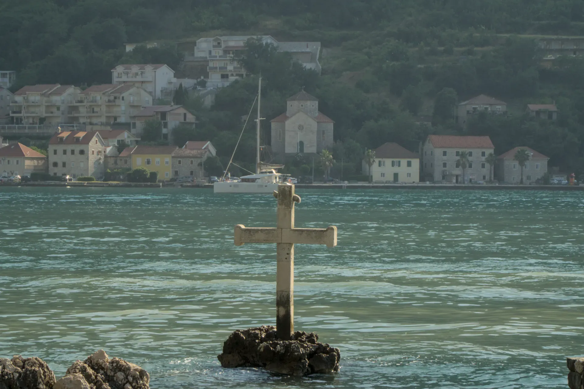 Une croix de pierre sur un rocher dans l'eau de la baie de Kotor, Monténégro, avec une ville côtière et des collines verdoyantes à l'arrière-plan.