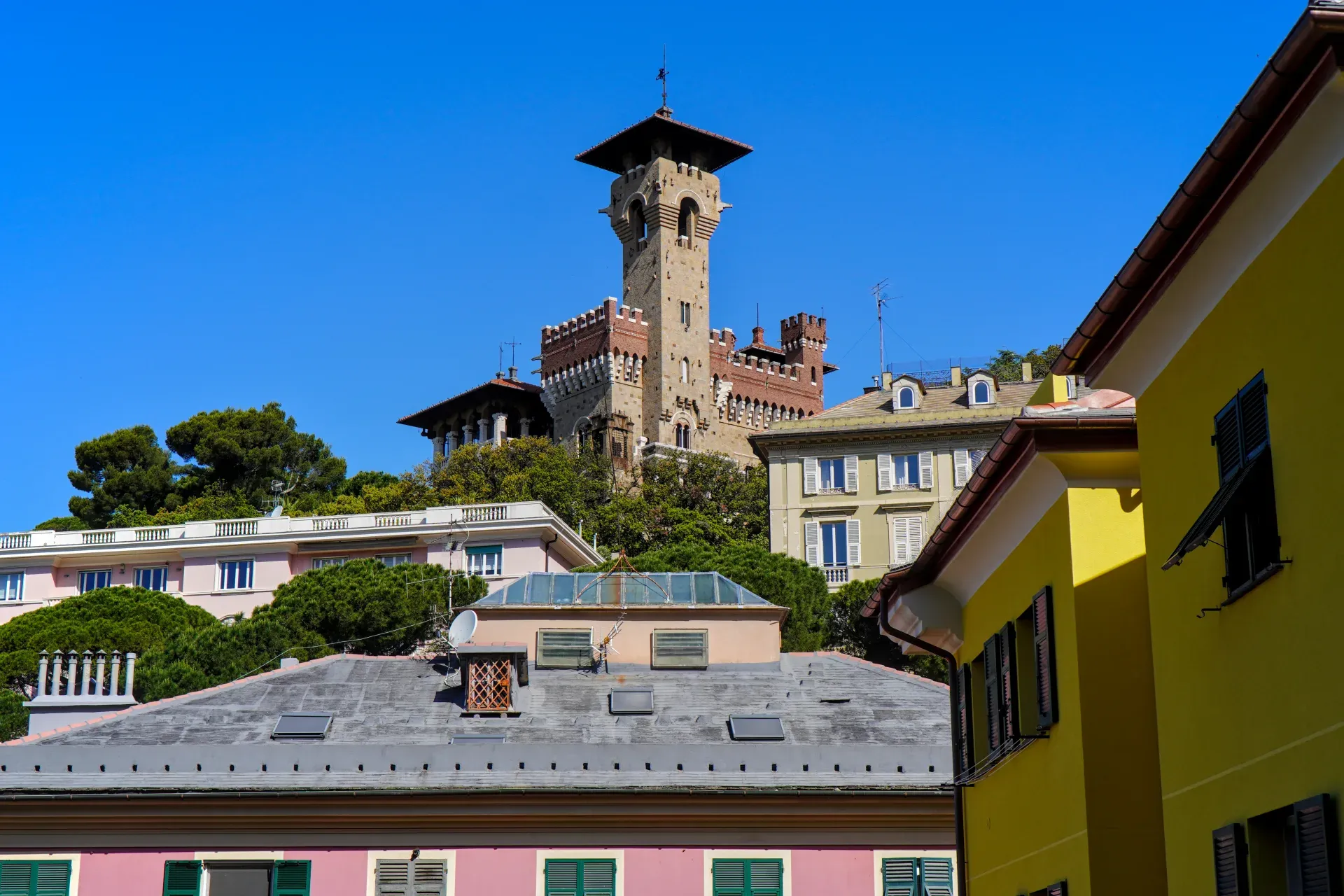 Una vista del Castello Mackenzie, un castello a Genova, Italia, con la sua torre imponente, sullo sfondo di alberi verdi ed edifici residenziali sotto un cielo blu brillante.