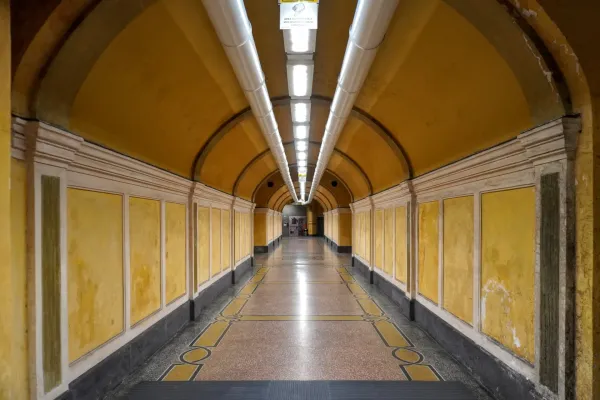 Un lungo tunnel simmetrico con pareti gialle, cornici bianche, pavimento in marmo a motivi e luci a soffitto luminose.