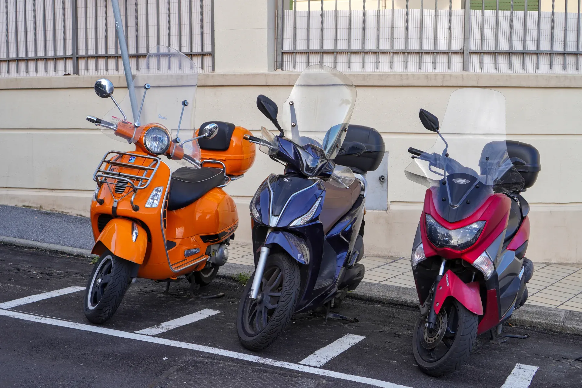 An orange Vespa scooter, a dark blue scooter, and a red scooter are parked in a row on a street with white parking lines.