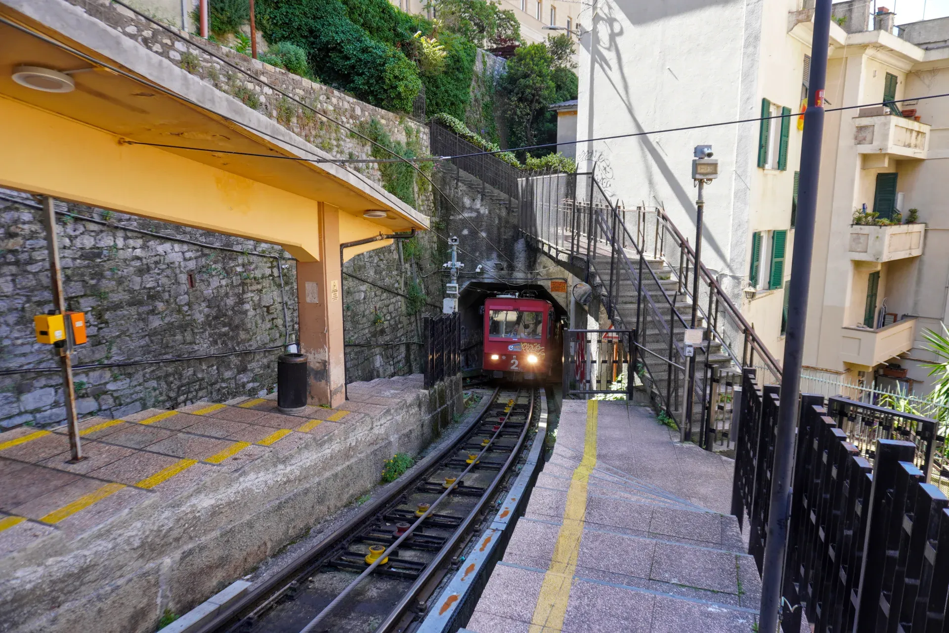 Crveni funi­kular sa upaljenim farovima ulazi u tunel na strmoj železničkoj pruzi. Pogled je sa perona stanice sa žutim krovom, naslonjenim na kameni zid. Desno, stepenište sa crnom ogradom penje se uzbrdo pored zgrade sa balkonima.