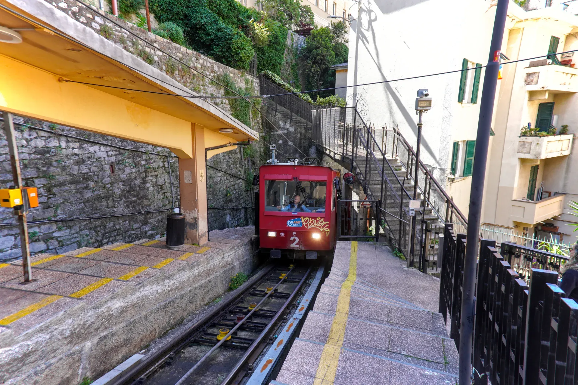 Un train funiculaire rouge portant le numéro 2 à l'avant monte sur une voie escarpée à Gênes, Italie, à côté d'un quai avec un toit jaune.