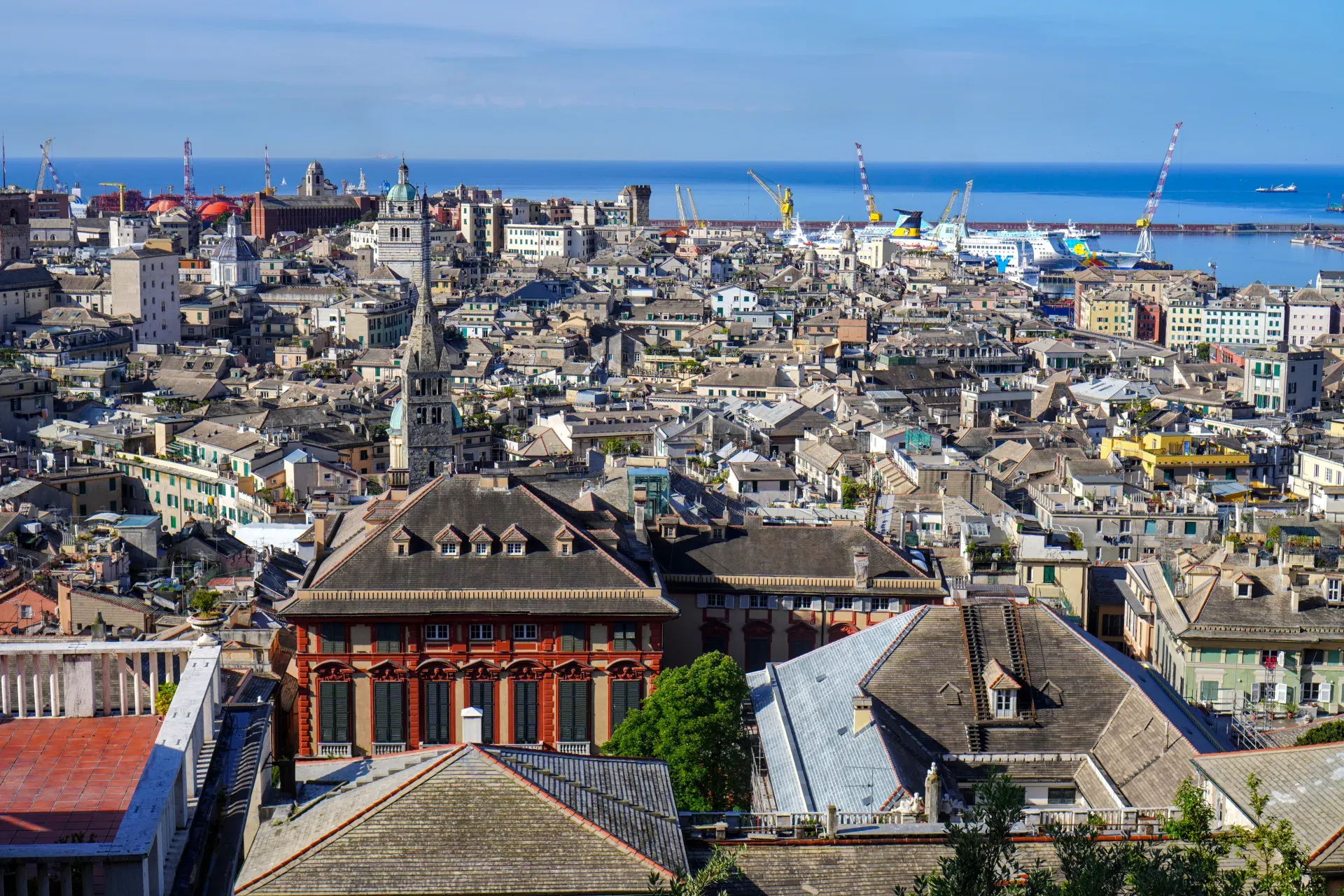 Una ripresa grandangolare di Genova, Italia, che mostra il porto attivo con navi cargo, traghetti e gru, affacciata sul denso paesaggio urbano che si estende verso il mare blu.