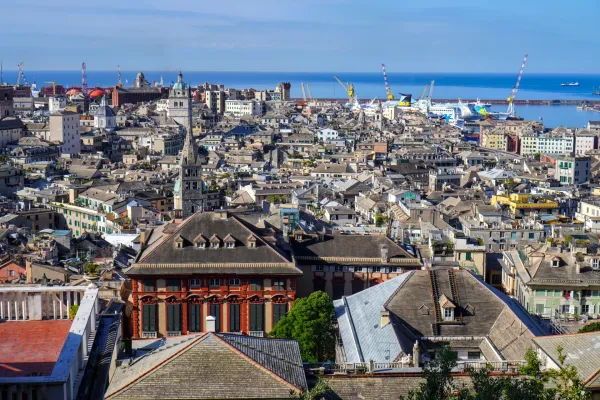 Una toma de gran angular de Génova, Italia, que muestra el puerto activo con buques de carga, ferries y grúas, con vistas al denso paisaje urbano que se extiende hacia el mar azul.