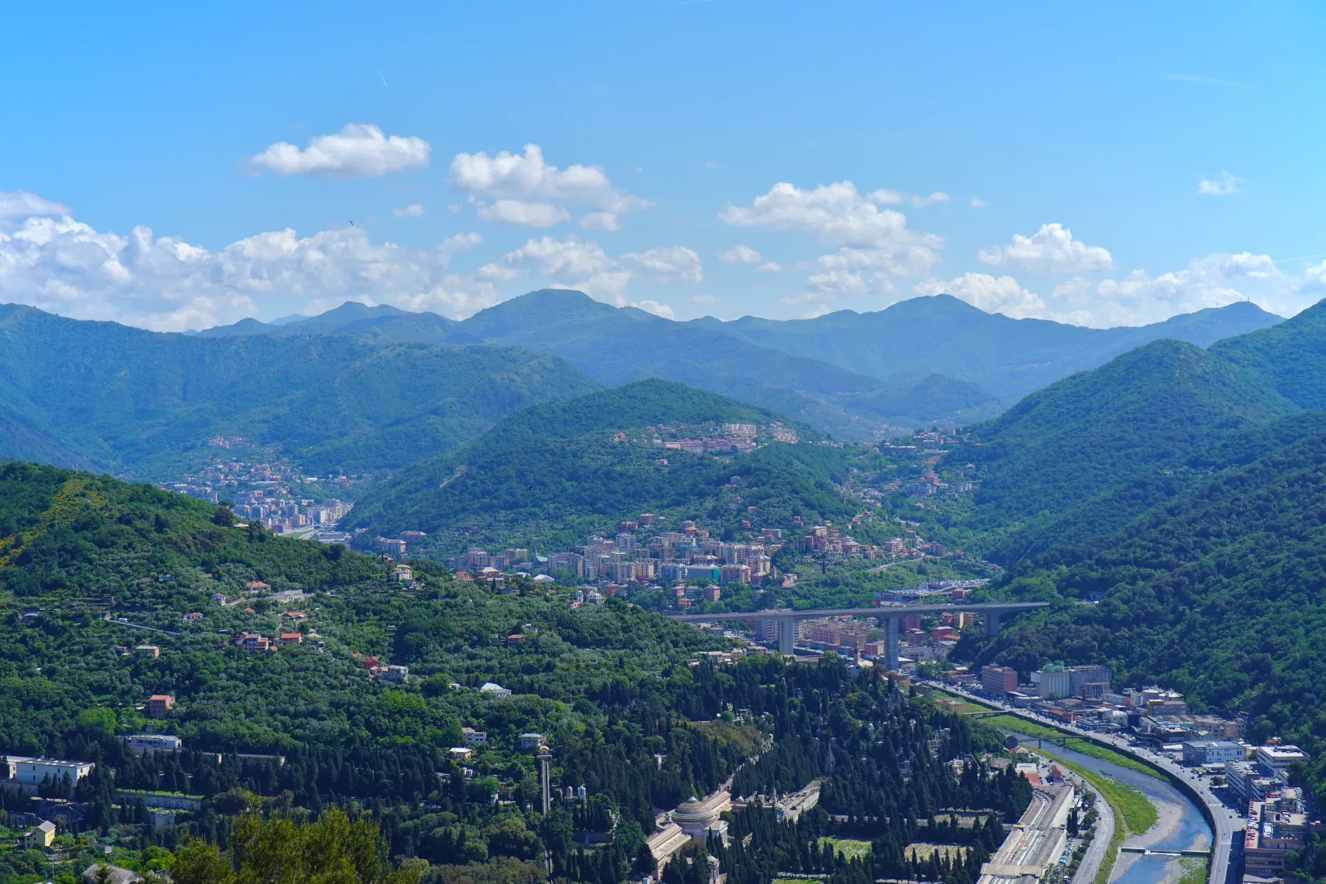 Vue sur des collines vertes couvertes d'arbres près de Gênes, Italie, avec d'anciennes fortifications et des tours de communication modernes sur les sommets, sous un ciel bleu vif.