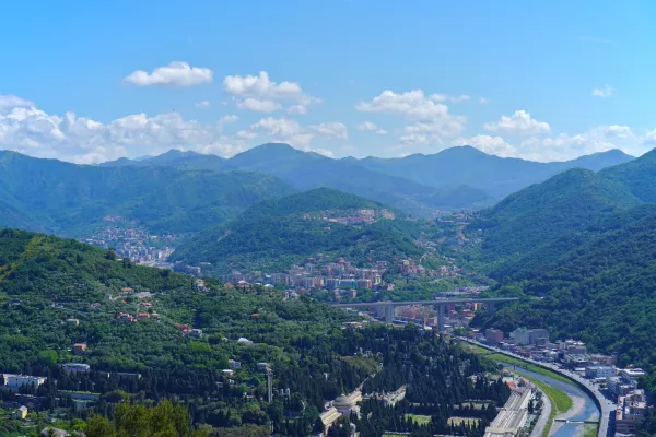 View of green, tree-covered hills near Genoa, Italy, with ancient fortifications and modern communication towers on the summits, under a bright blue sky.
