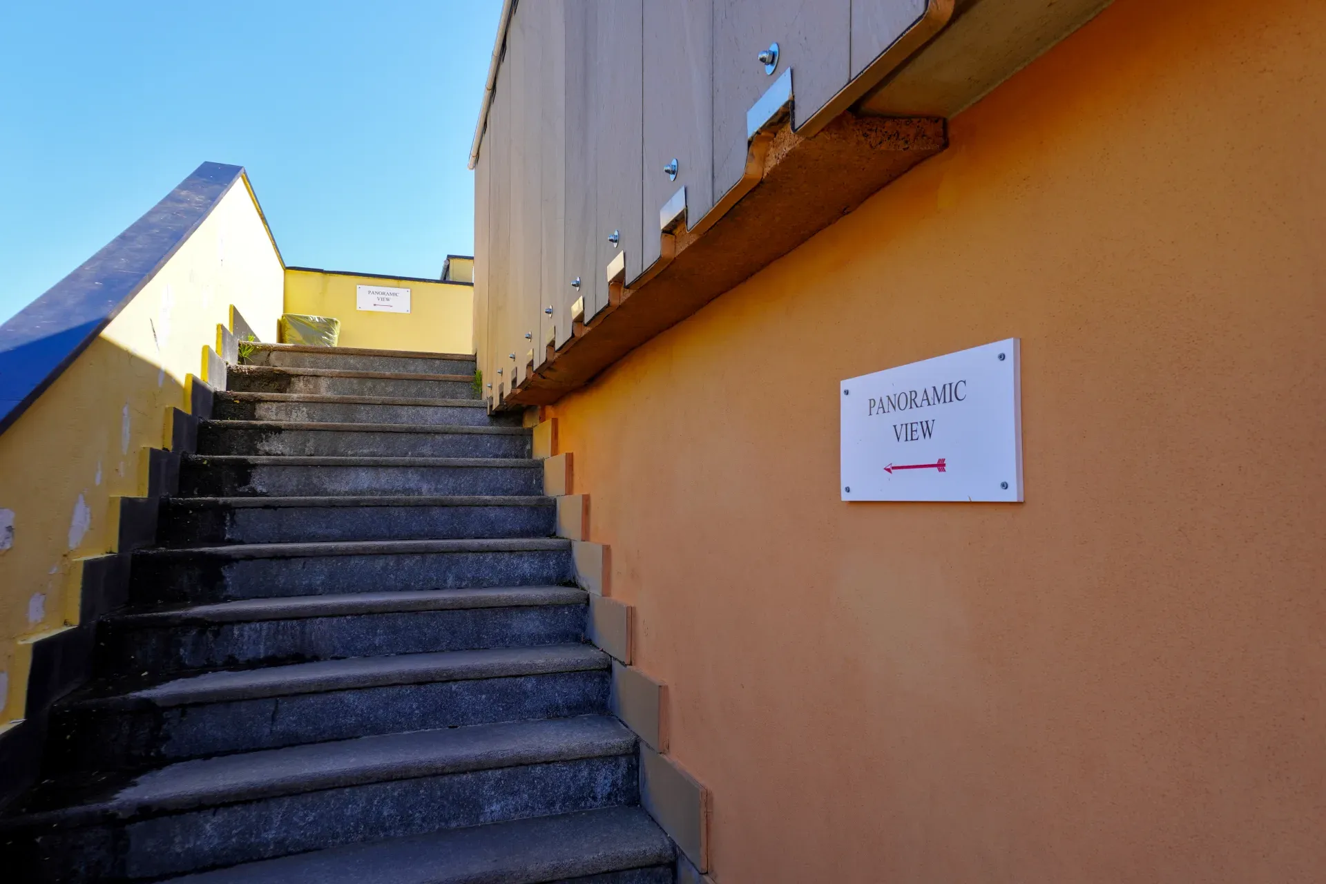 Una scala in pietra conduce a un cartello con la scritta 'PANORAMIC VIEW' e una freccia rossa che punta a sinistra, accanto a un muro colorato sotto un cielo blu limpido a Genova, Italia.
