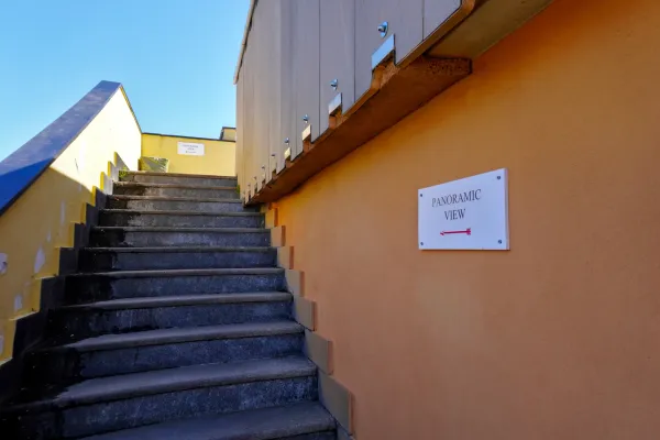 Un escalier en pierre mène à une pancarte indiquant 'PANORAMIC VIEW' avec une flèche rouge pointant vers la gauche, à côté d'un mur coloré sous un ciel bleu clair à Gênes, Italie.