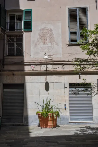 A sunlit narrow street in Genoa, Italy, with buildings featuring green and blue shutters, a faded fresco on the wall depicting a religious scene, a single street lamp, a large terracotta pot with plants, and two shop roll-up doors casting shadows on the paved ground.