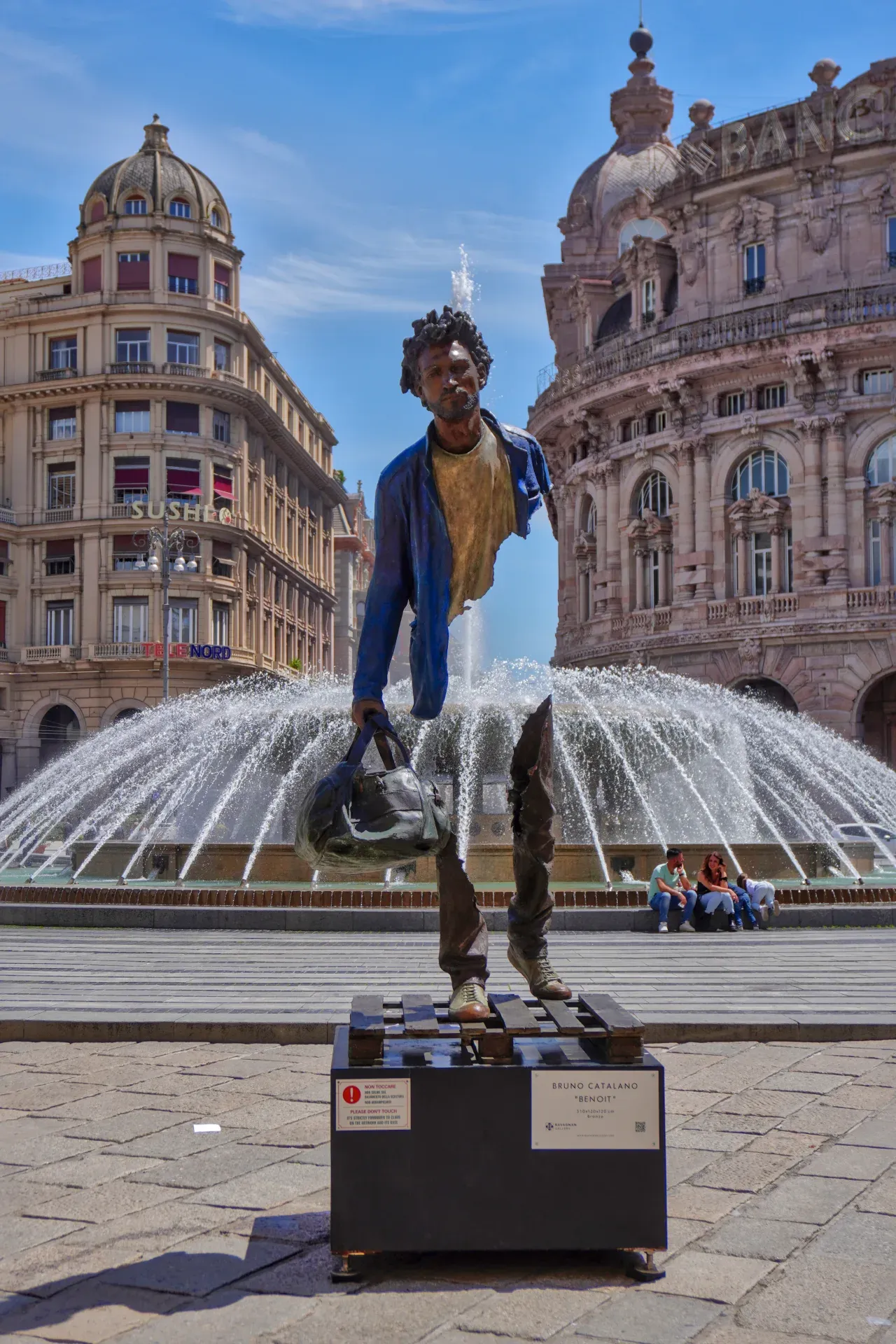 Une sculpture fragmentée en bronze d'un homme portant un sac, intitulée 'Benoit' par Bruno Catalano, se dresse devant une grande fontaine sur une place de ville à Gênes, Italie. Des bâtiments historiques sont visibles à l'arrière-plan, et des personnes sont assises près de la fontaine.