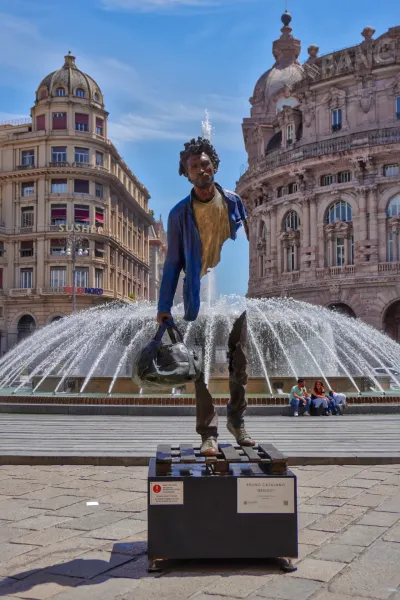 Fragmentovana bronzana skulptura muškarca koji nosi torbu, pod nazivom 'Benoit' Brune Catalana, stoji ispred velikog fontana na gradskom trgu u Đenovi, Italija. Istorijske zgrade su vidljive u pozadini, a ljudi sede pored fontana.
