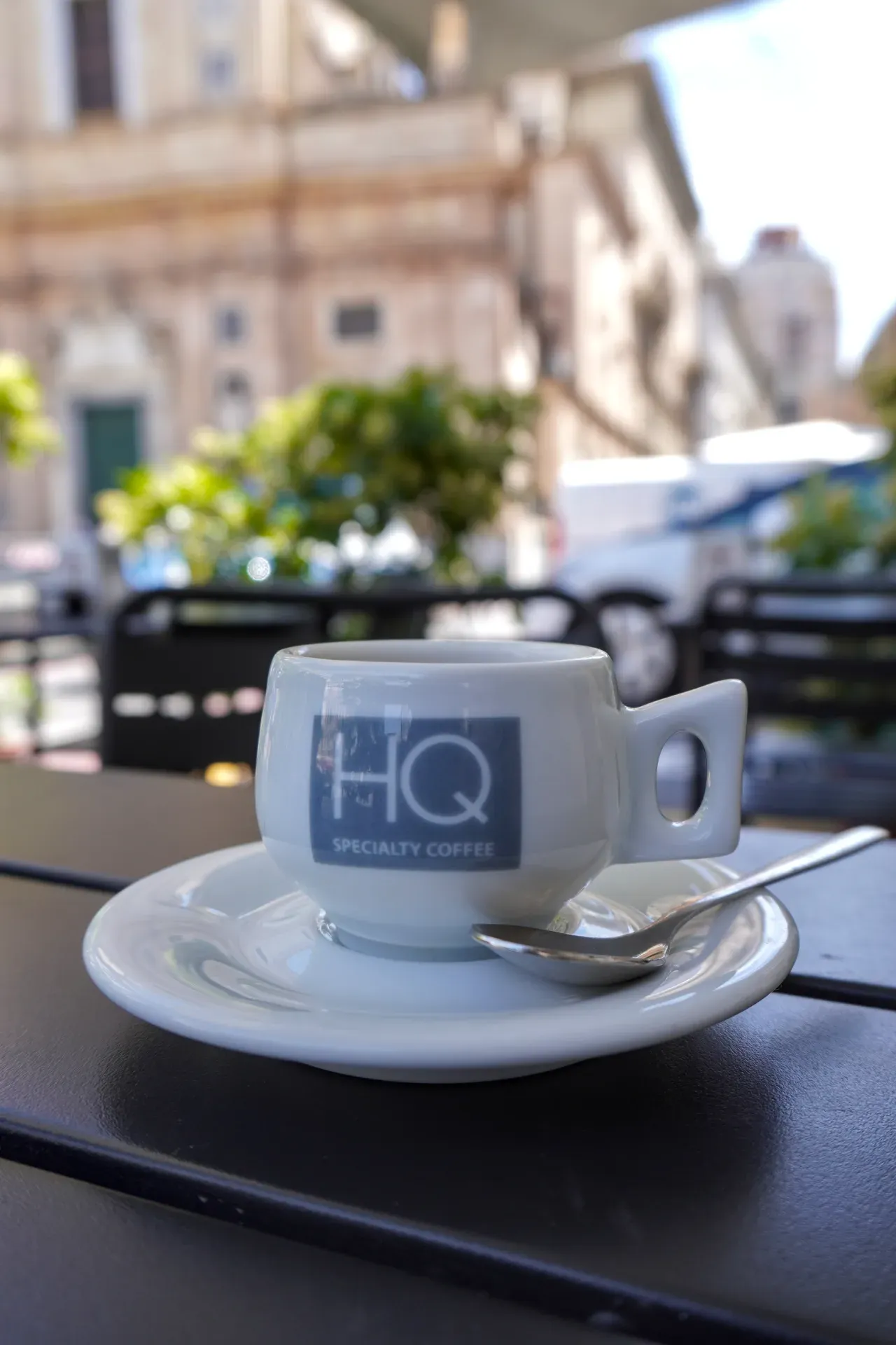 Eine weiße Espressotasse mit dem 'HQ Specialty Coffee'-Logo und einem Löffel auf einer Untertasse, auf einem schwarzen Tisch in einem Café im Freien in Genua, Italien. Der Hintergrund ist unscharf mit Gebäuden und Grünflächen.