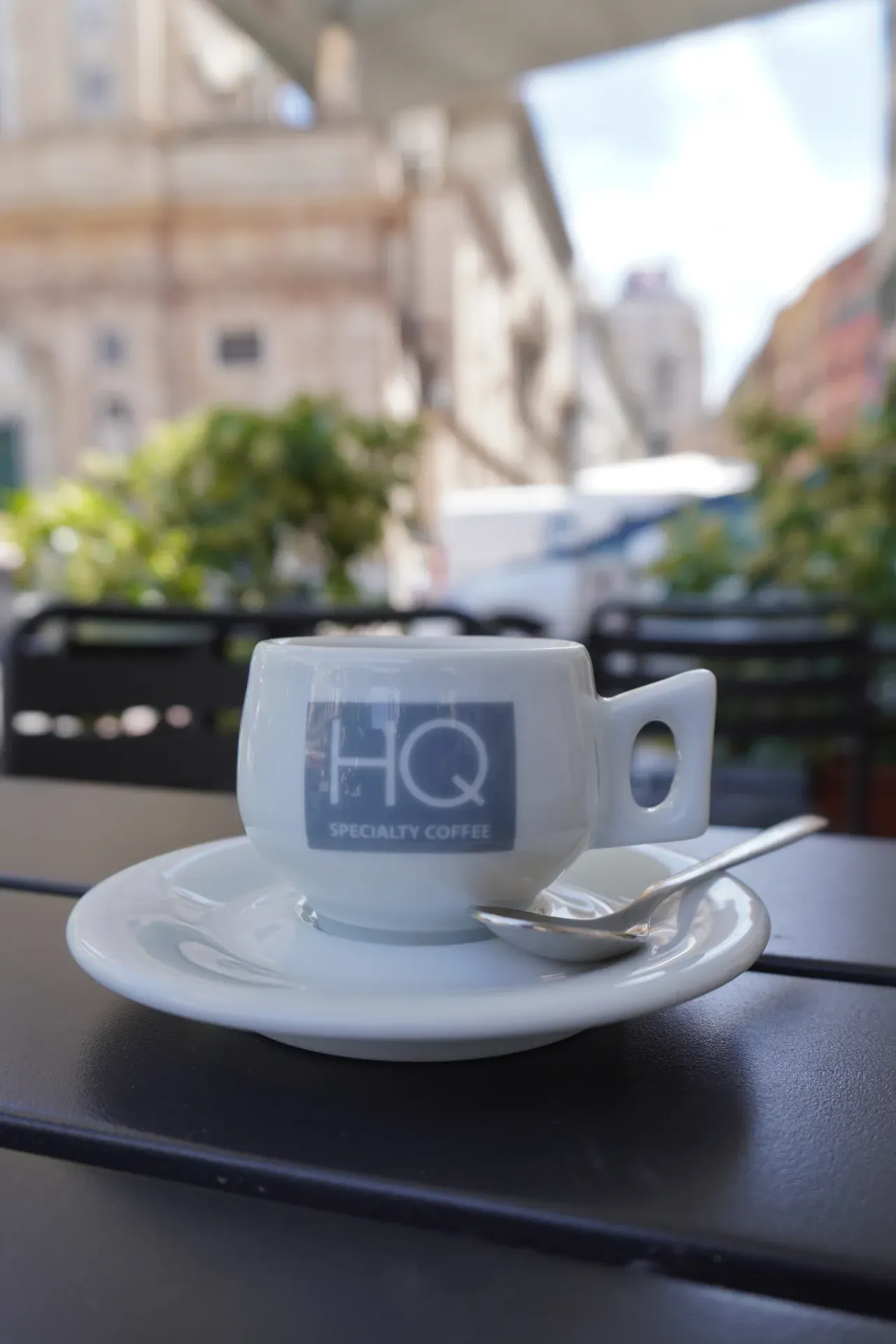 Белая чашка для эспрессо с логотипом 'HQ Specialty Coffee' стоит на блюдце с ложкой на столике на открытом воздухе в Генуе, Италия, на размытом фоне зданий и зелени.
