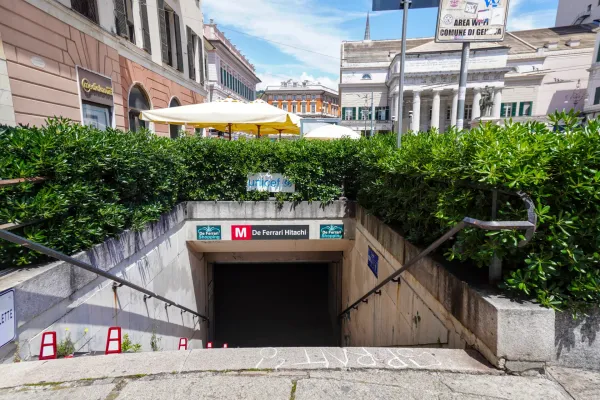 Pogled sa ulice na ulaz u stanicu metroa De Ferrari u Đenovi, Italija, sa zelenim živicama, zgradama i natpisima.