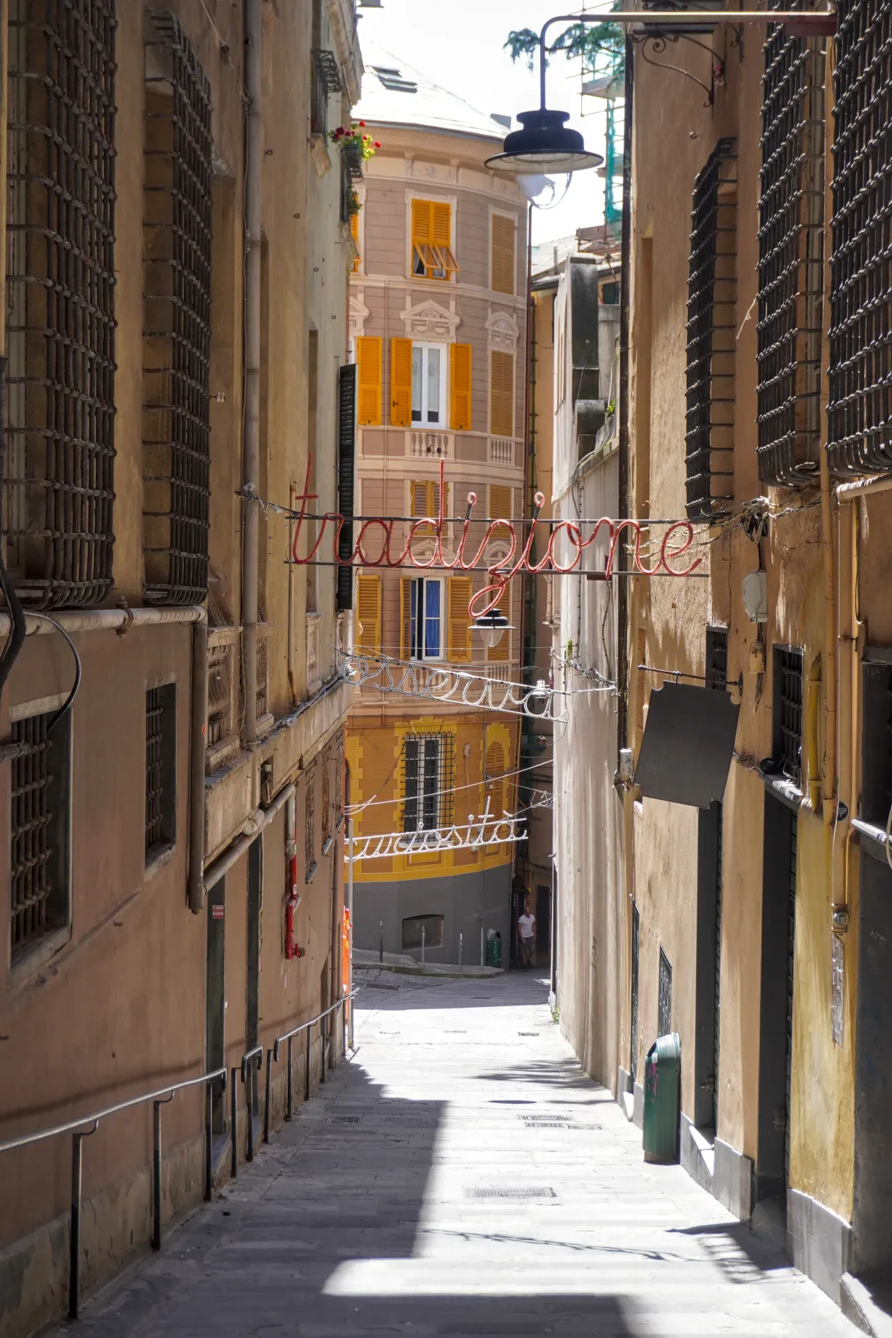 Una calle estrecha en Génova, Italia, con edificios altos a ambos lados y letreros de neón que dicen 'Tradizione' y 'Curiosità' colgando sobre el pavimento de adoquines.
