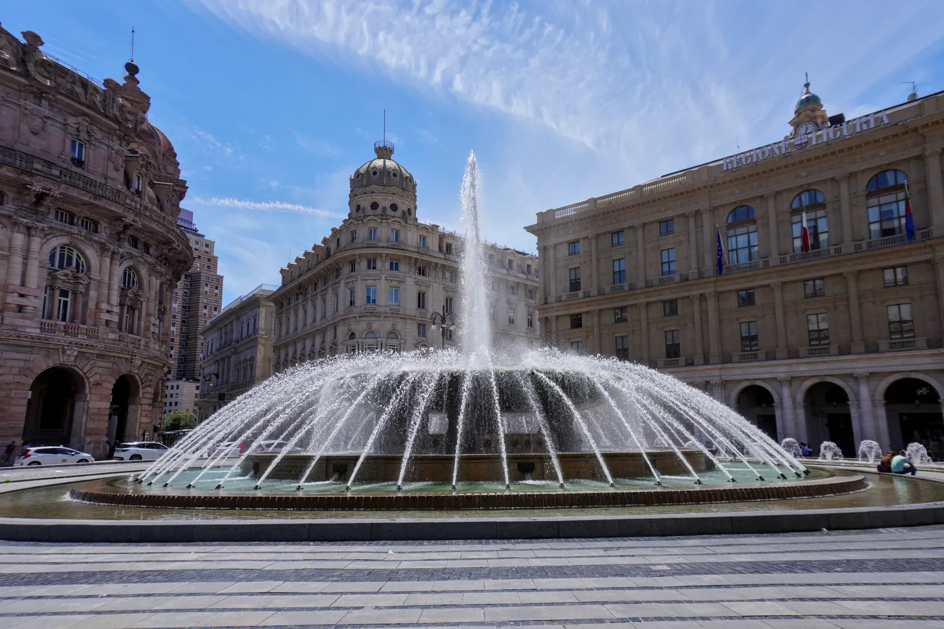 A large, circular fountain sprays water in the center of a square, surrounded by elegant historic buildings in Genoa, Italy. The sky above is blue with dramatic wispy clouds.