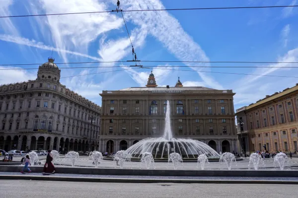 A large, multi-tiered fountain sprays water in Piazza De Ferrari, Genoa, Italy. In the foreground, white cars, one a taxi, and a cyclist are on the road. Behind the fountain is a grand yellow building with pink accents under a blue sky with white clouds.