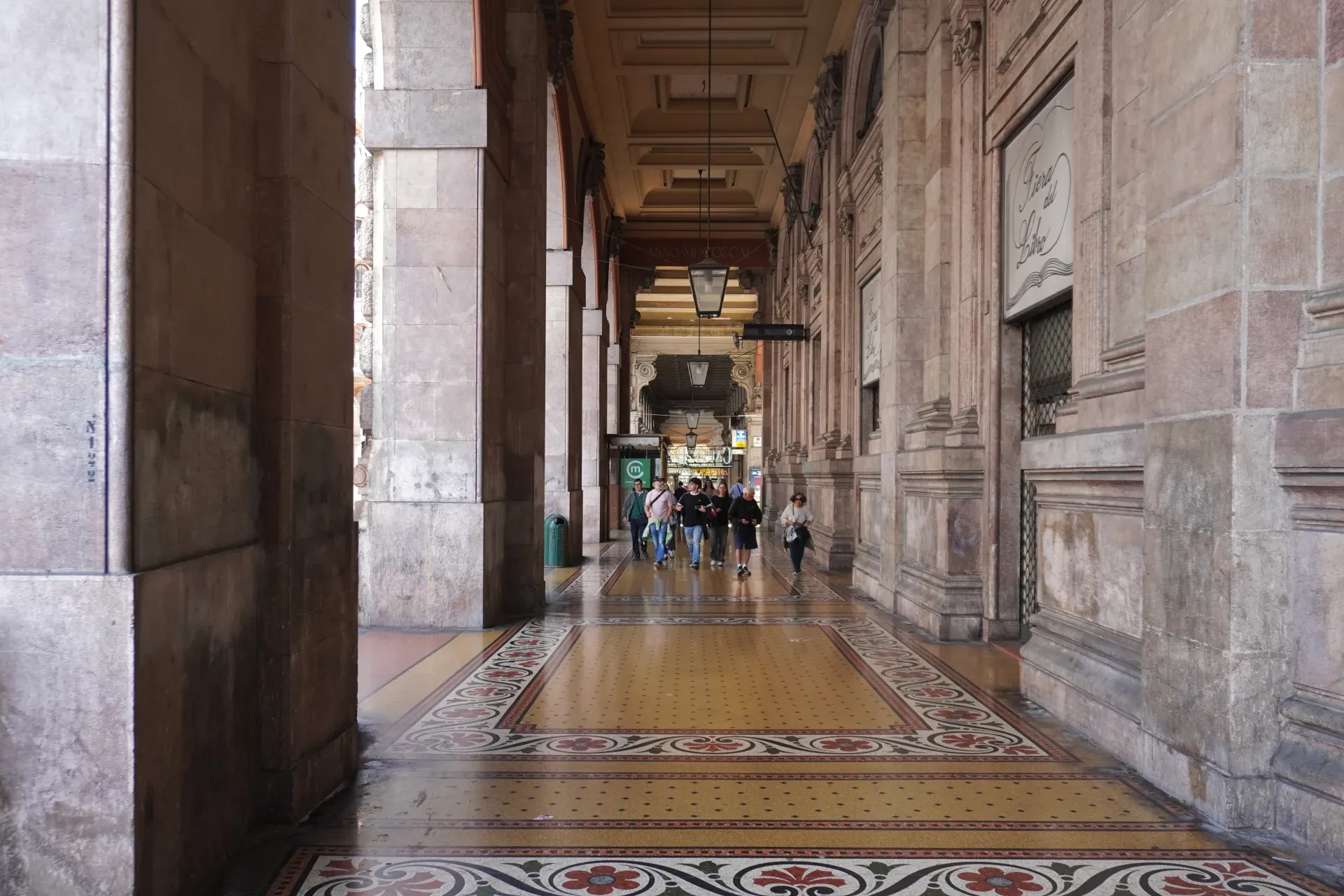 Une perspective d'une allée couverte à Gênes, Italie, avec de grandes arches en pierre, un escalier en marbre et un sol coloré en carreaux de mosaïque. La lumière du soleil projette des ombres sur le sol, illuminant les décorations ornées.