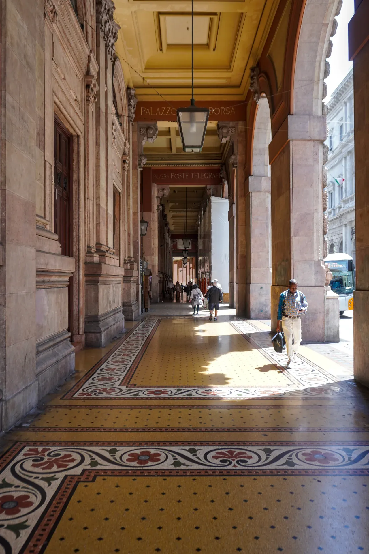 A person walks through a wide, covered walkway with tall arches, ornate columns, and a patterned tiled floor in Genoa, Italy.