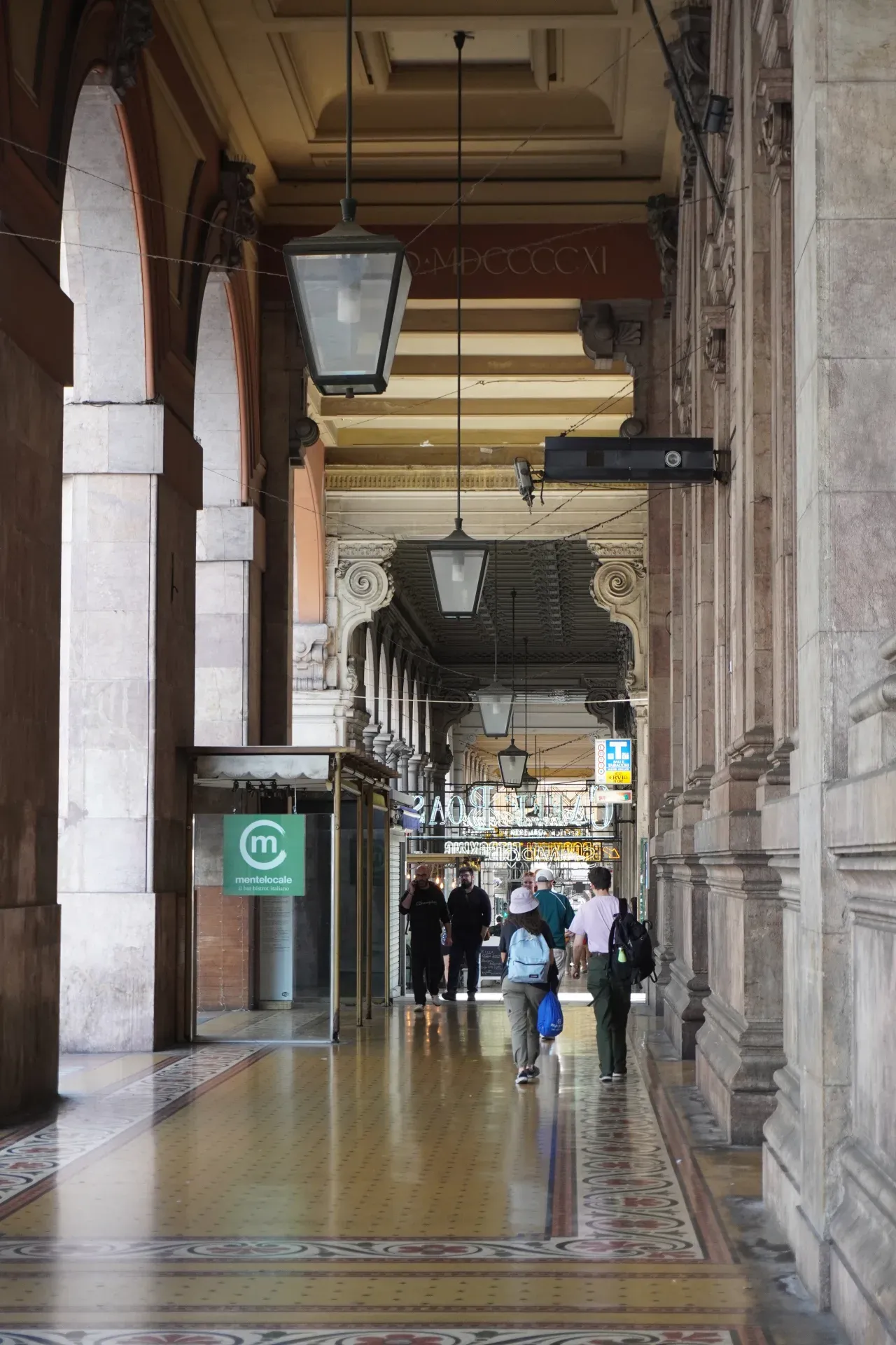Une longue allée couverte aux colonnes cintrées mène au Palazzo della Borsa. Le sol est orné de carreaux de mosaïque colorés, et la lumière du soleil y pénètre, projetant des ombres. Plusieurs personnes s'éloignent du spectateur le long du portique.
